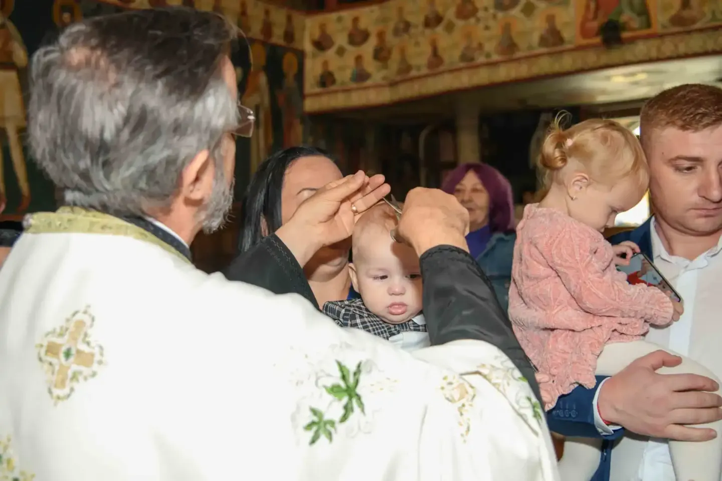 Priester segnet Säugling bei Taufzeremonie, Familie beobachtet - Eventfotografie Wien