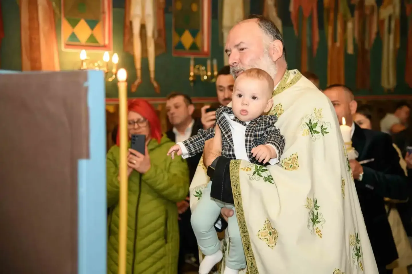 Priester in weißem Ornat hält Kleinkind während Taufzeremonie in Kirche, Eventfotograf Wien dokumentiert Moment