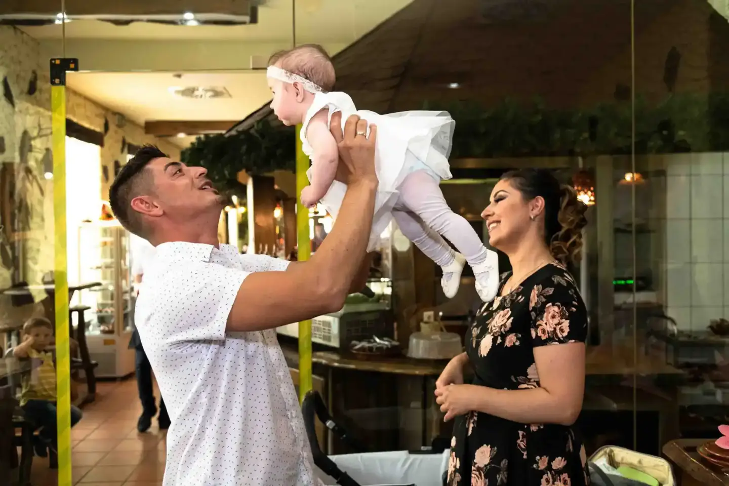 Vater hebt lachendes Baby in die Luft, Mutter schaut lächelnd zu – professionelle Eventfotografie einer Taufe in Wien