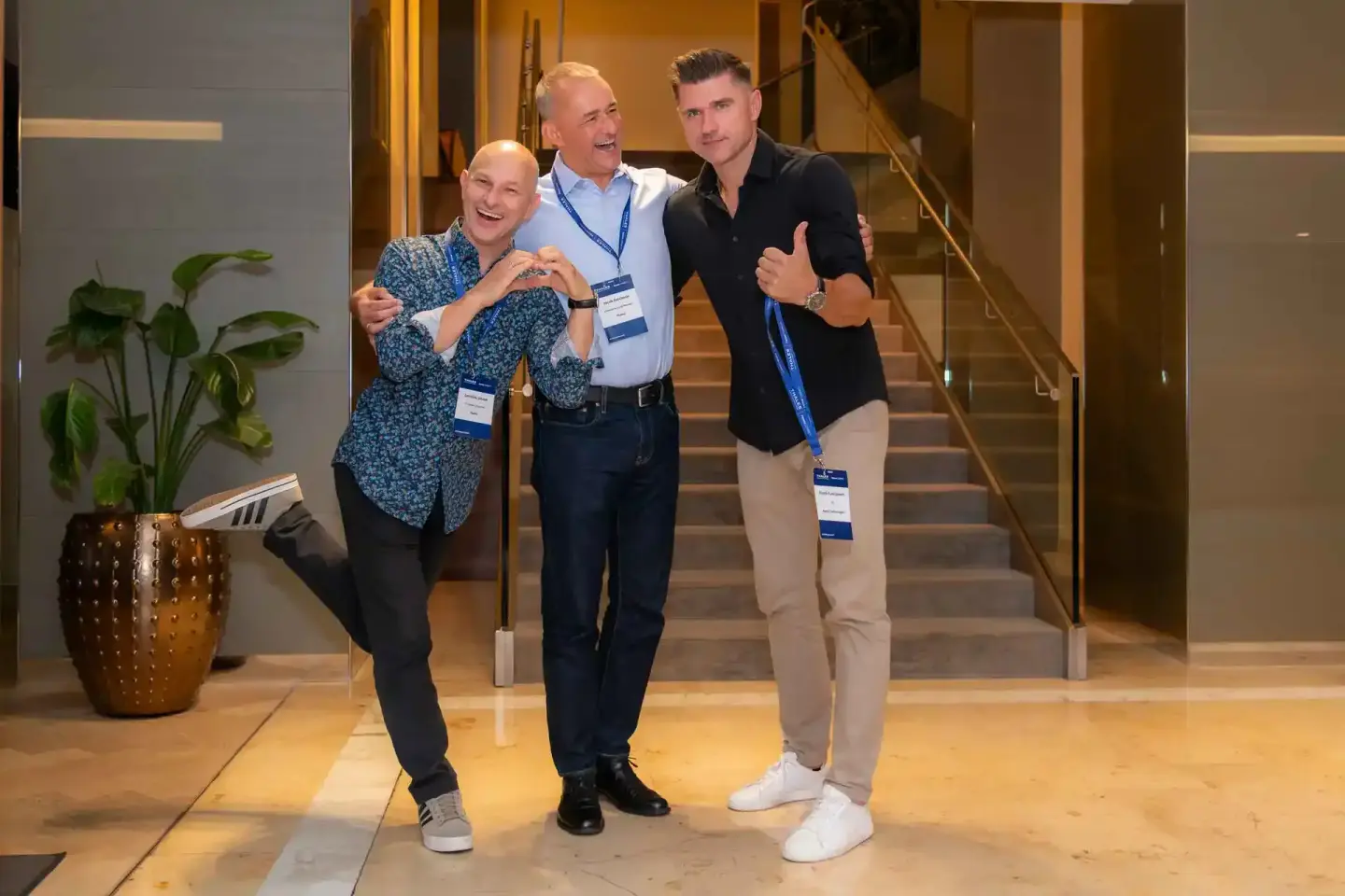 Drei Männer lachen zusammen bei einem Firmenevent Wien, tragen Ausweisbänder und posieren vor einer modernen Treppe