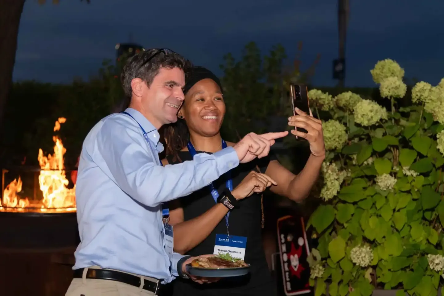 Zwei Personen mit Namensschildern nehmen ein Selfie beim Thales Firmenevent Wien auf, im Hintergrund lodert ein Lagerfeuer