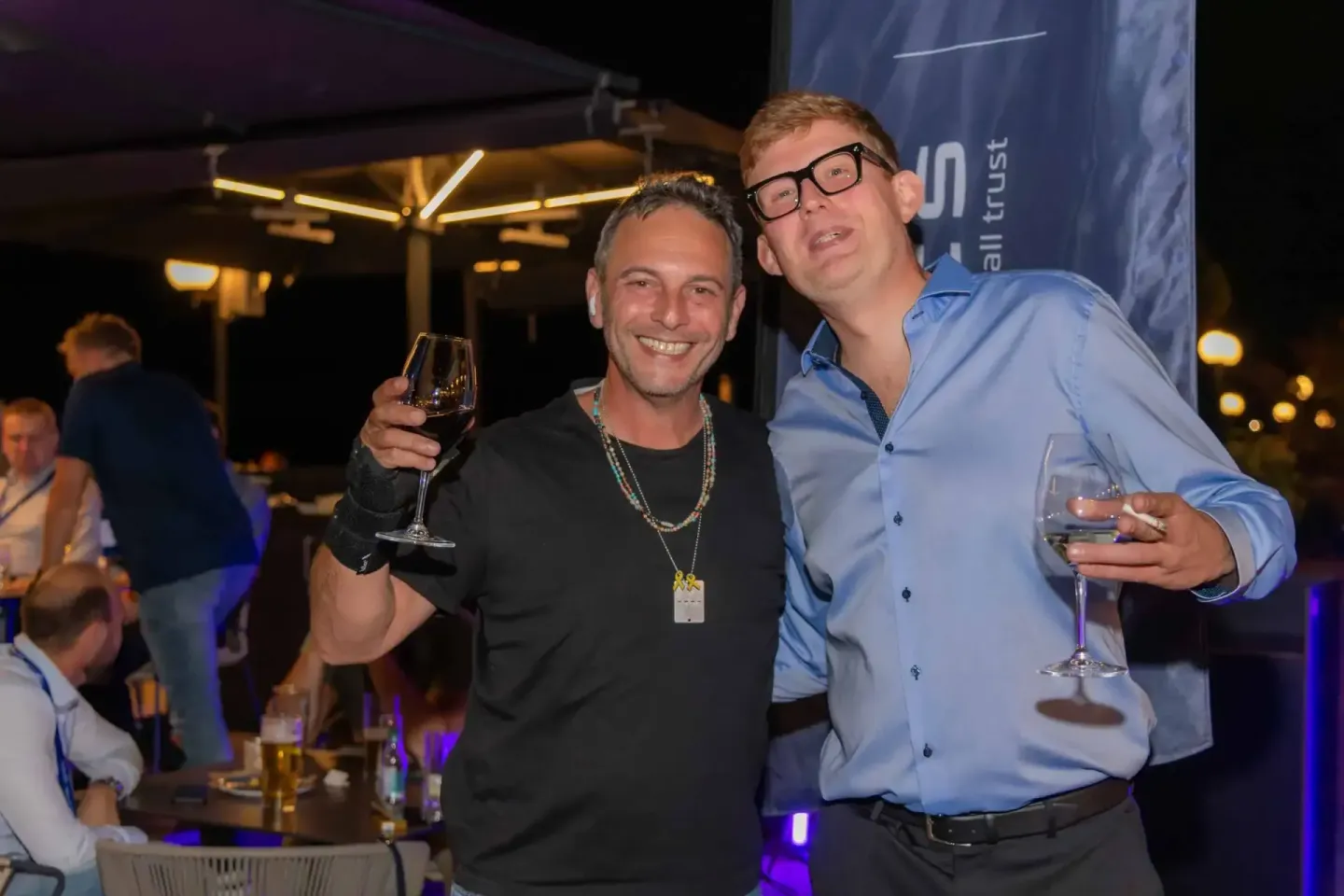 Zwei Männer stoßen mit Weingläsern an einem Firmenevent Wien an, im Hintergrund weitere Gäste unter einer Terrassenüberdachun