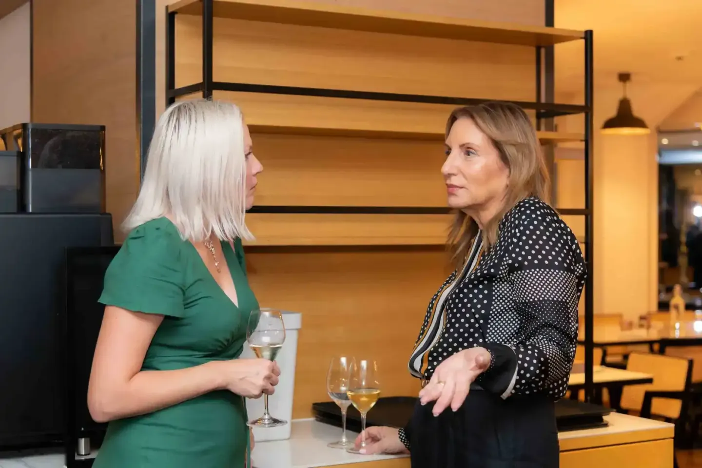 Zwei Frauen im eleganten Gespräch bei einem Firmenevent in Wien, eine trägt grünes Kleid, die andere schwarzes Polka-Dot-Blaz