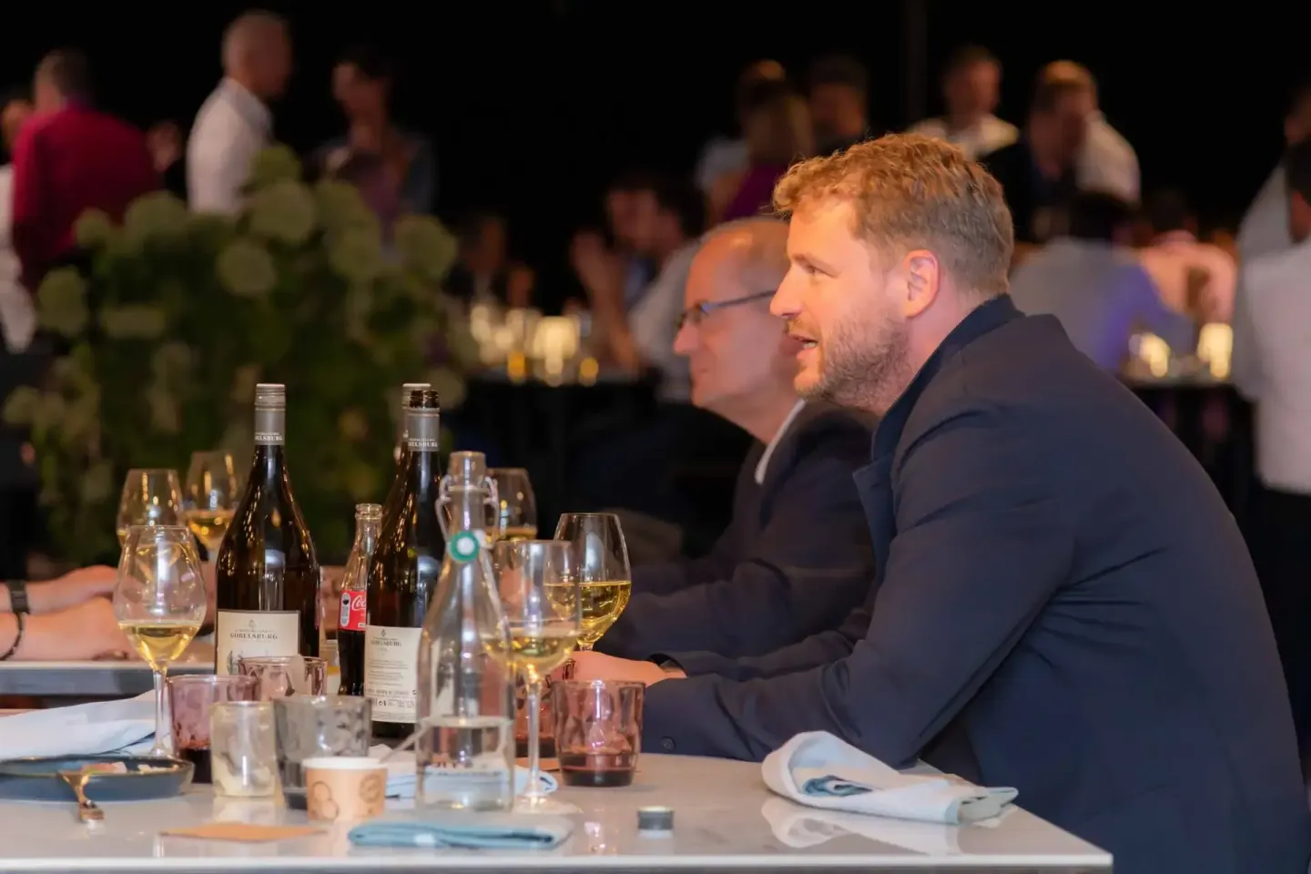 Zwei Geschäftsmänner sitzen an einem gedeckten Tisch mit Weingläsern und Weinflaschen bei einem Firmenevent in Wien