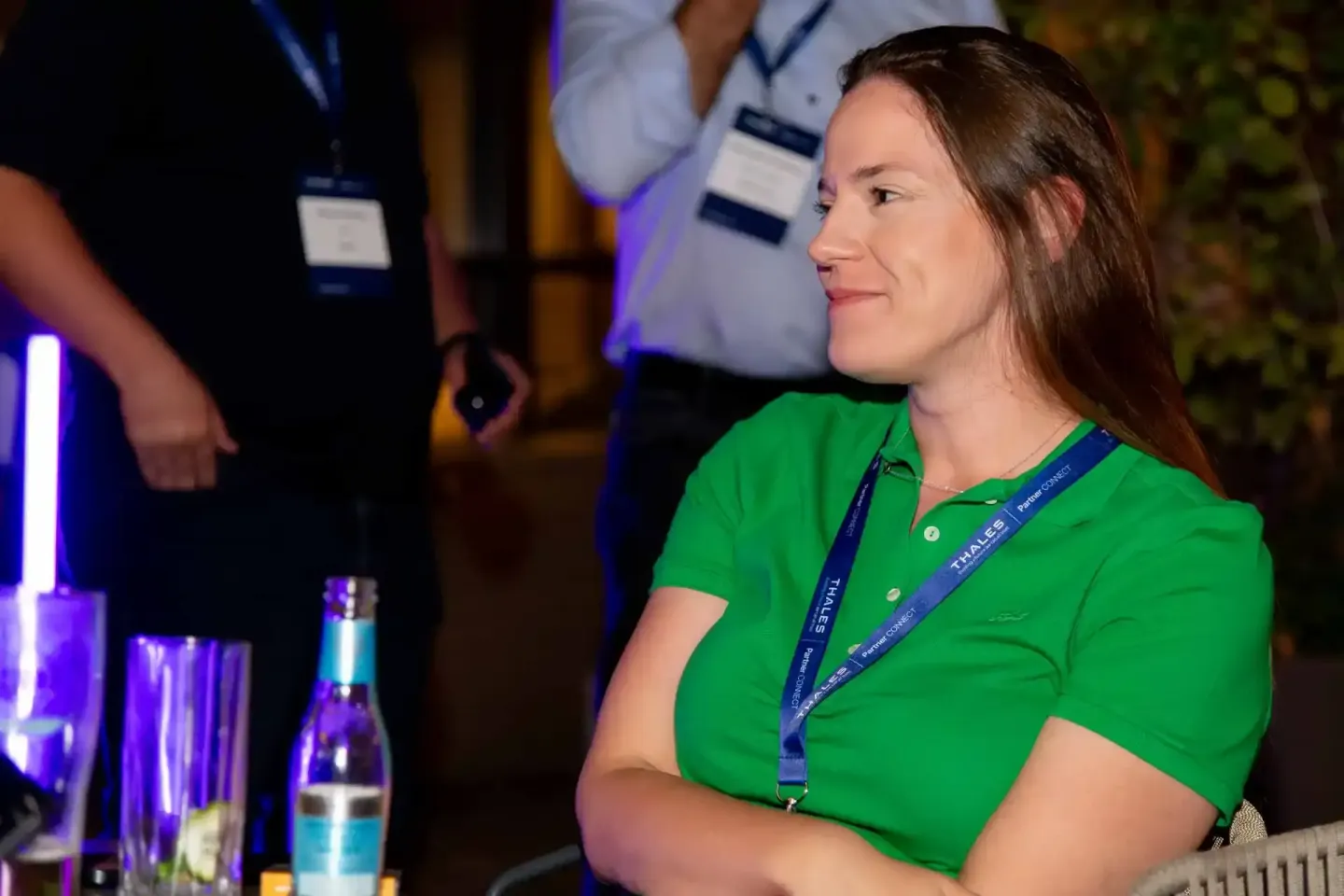 Frau in grünem Polo-Shirt mit Lanyard bei Thales Firmenevent Wien, sitzt am Tisch mit Getränken