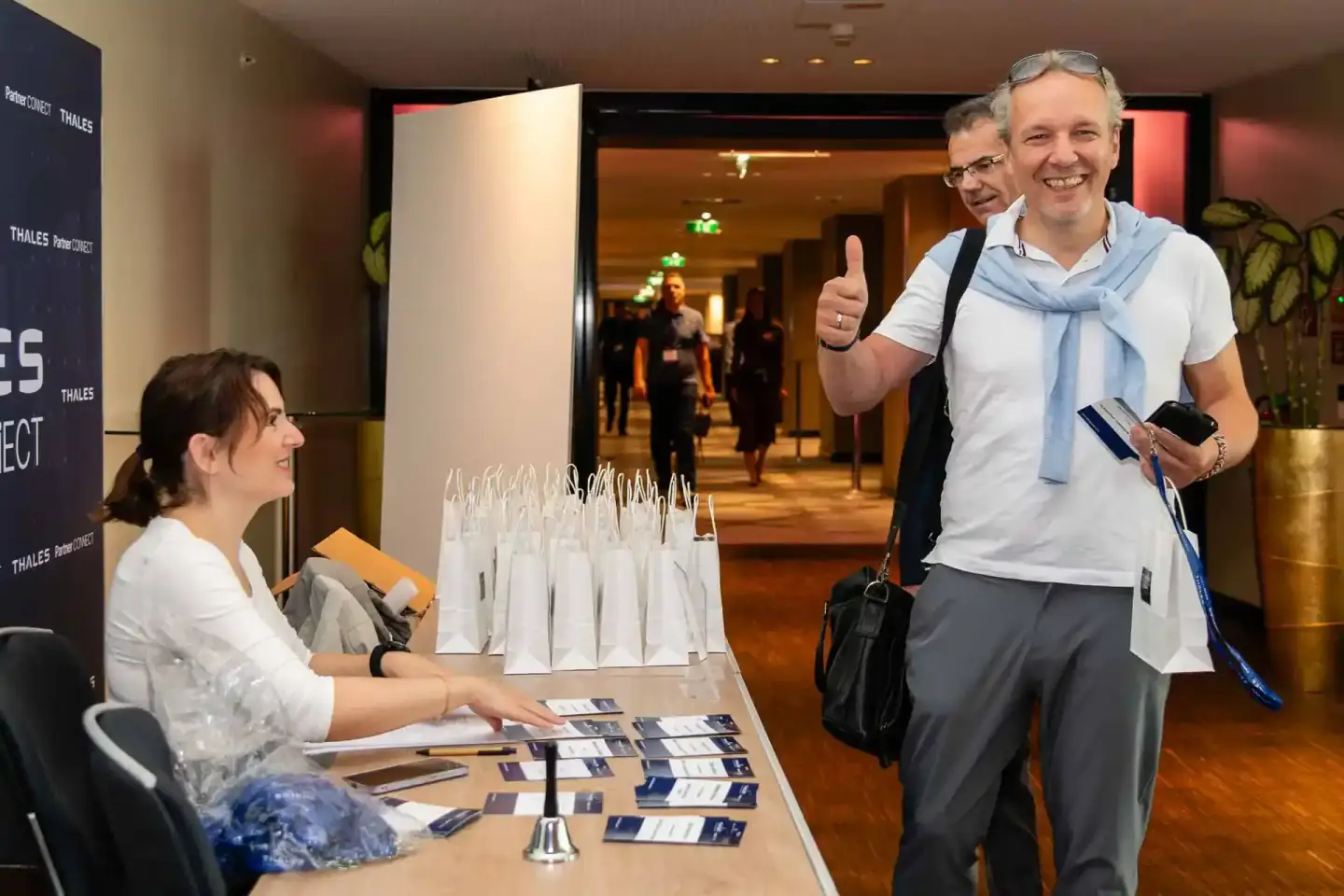 Eventfotograf dokumentiert Thales Firmenevent in Wien: Mann mit Daumen hoch an Registrierungstisch, Besucherin hinter Counter