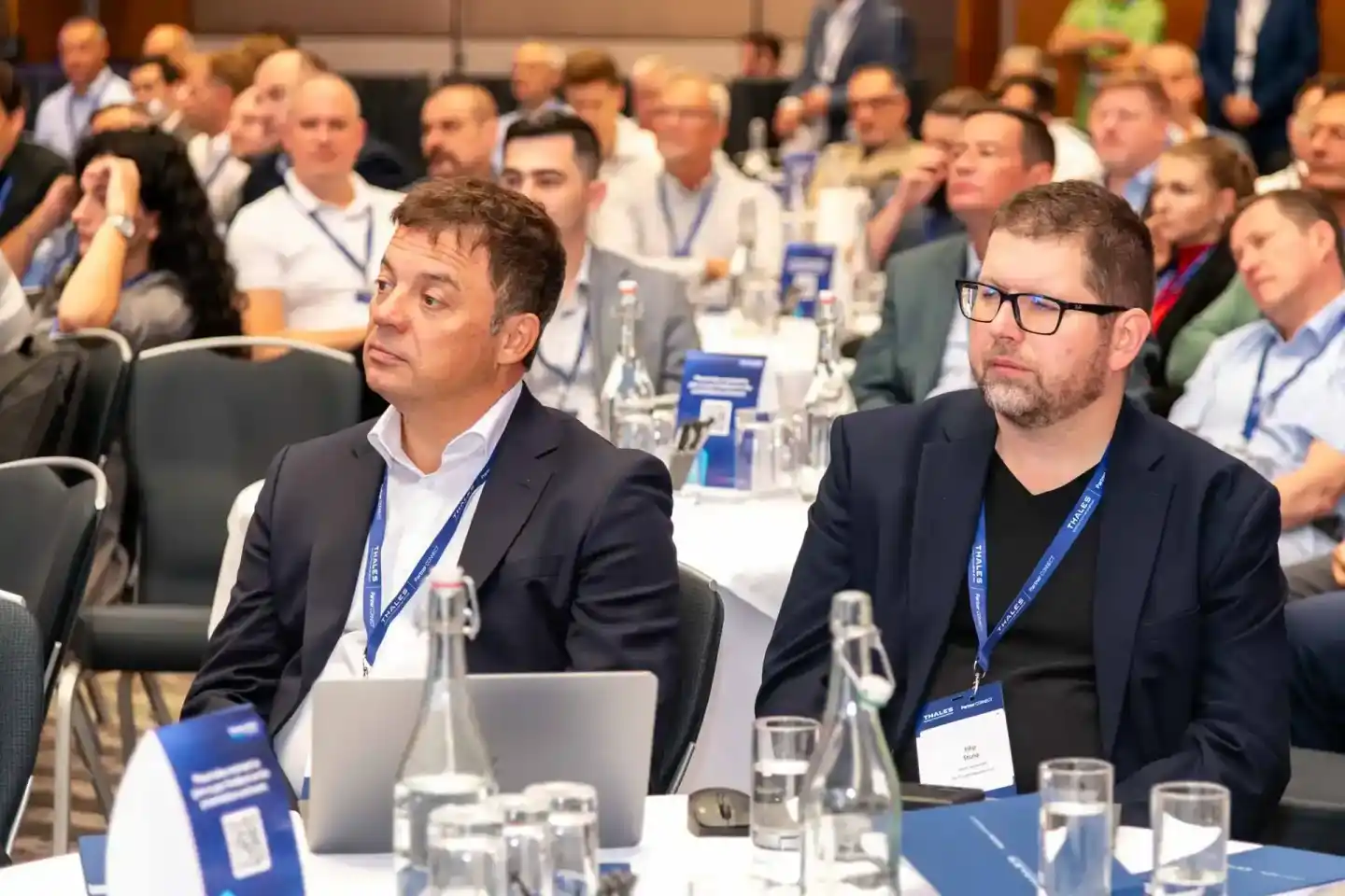 Zwei Geschäftsmänner mit Konferenzausweisen sitzen an einem Tisch während einer Geschäftsveranstaltung in Wien, im Hintergrun