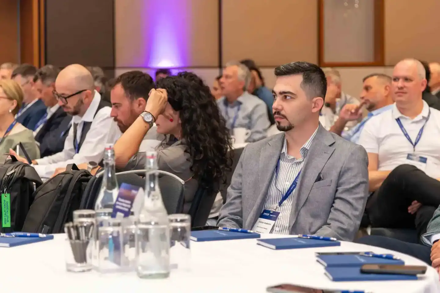 Teilnehmer eines Firmenevent Wien sitzen aufmerksam bei einer Konferenz, vorne ein Mann im grauen Anzug mit Namensschild