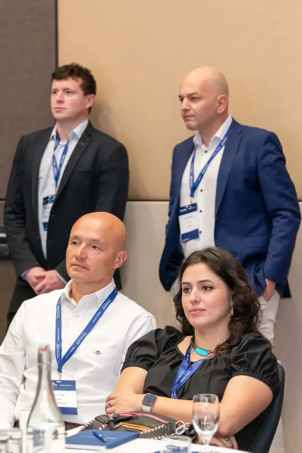 Vier Geschäftspersonen bei einem Firmenevent Wien mit Namensschildern, zwei stehend im Hintergrund, zwei sitzend am Tisch