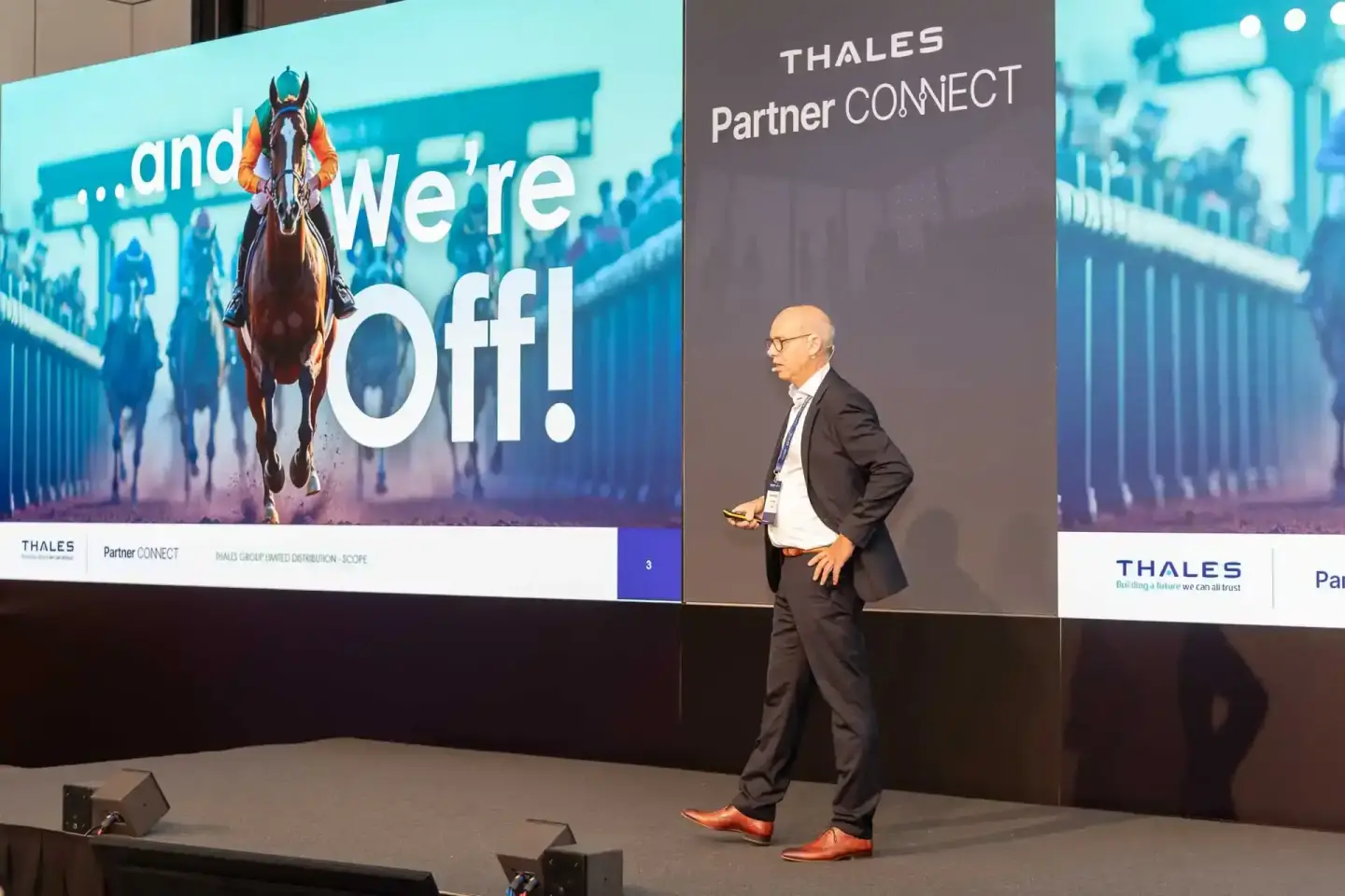 Geschäftsmann präsentiert auf Thales Partner Connect Veranstaltung vor großer Leinwand mit Pferderennen-Motiv - Eventfotograf