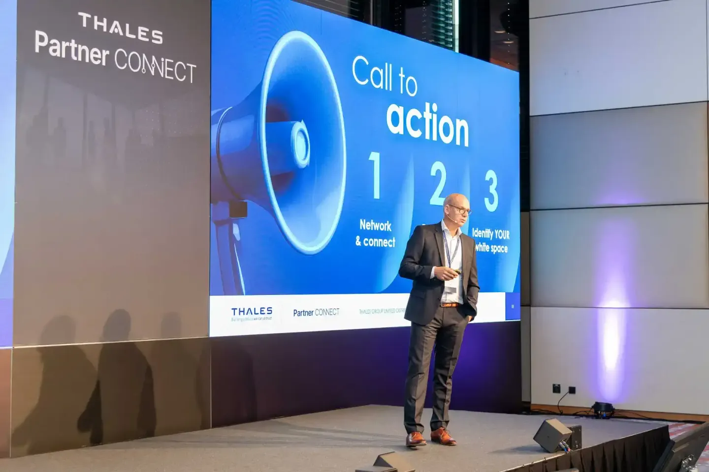 Geschäftsmann präsentiert auf der Thales Partner Connect Konferenz mit Megafon-Grafik und Call-to-Action auf großer Bühne – E