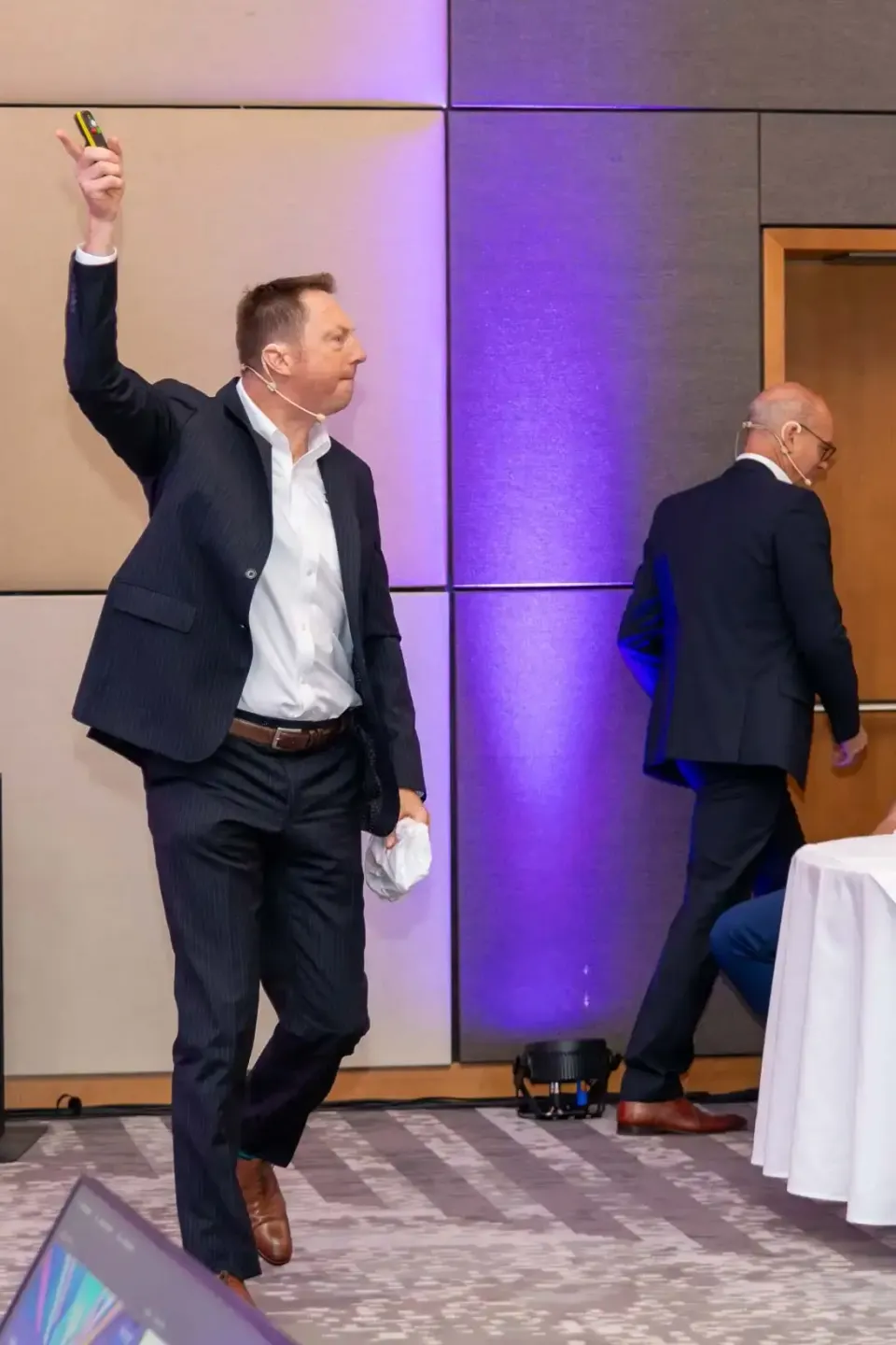 Geschäftsmann in dunklem Anzug mit erhobener Faust auf einem Firmenevent in Wien, im Hintergrund ein weiterer Mann und modern