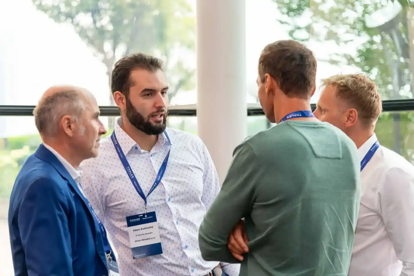 Vier Geschäftsmänner in Businesskleidung mit Namensschildern diskutieren auf einer Firmenveranstaltung in Wien vor großen Fen