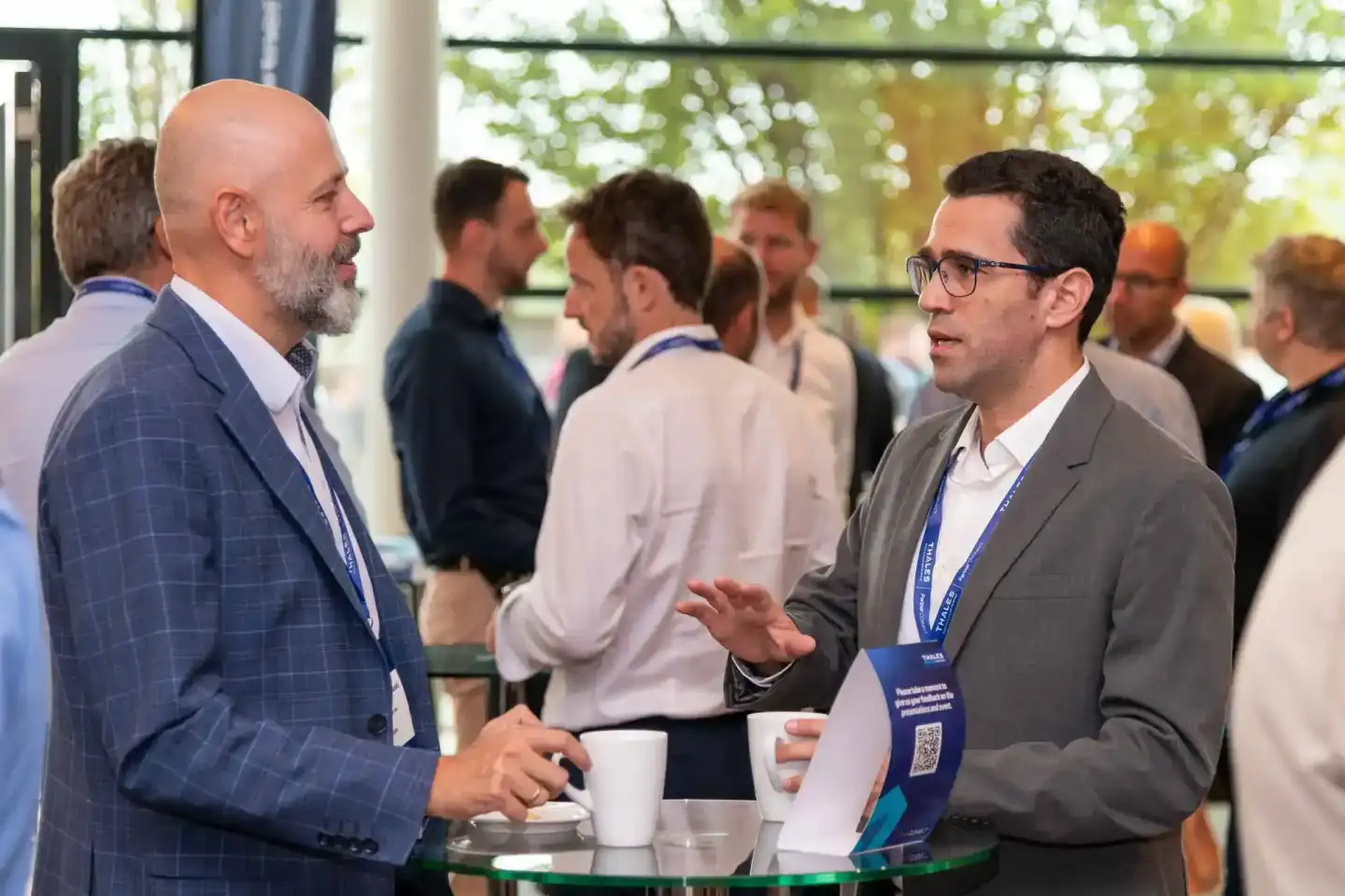 Zwei Geschäftsmänner in Anzügen mit Namensausweisen im Gespräch auf einem Firmenevent Wien, weitere Teilnehmer im Hintergrund