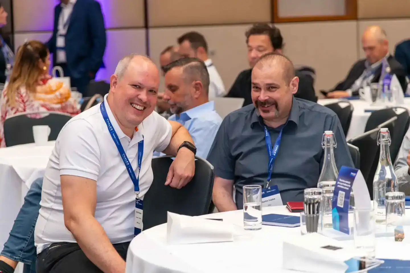 Zwei lachende Männer mit Lanyards sitzen an einem Konferenztisch beim Thales Firmenevent Wien und unterhalten sich