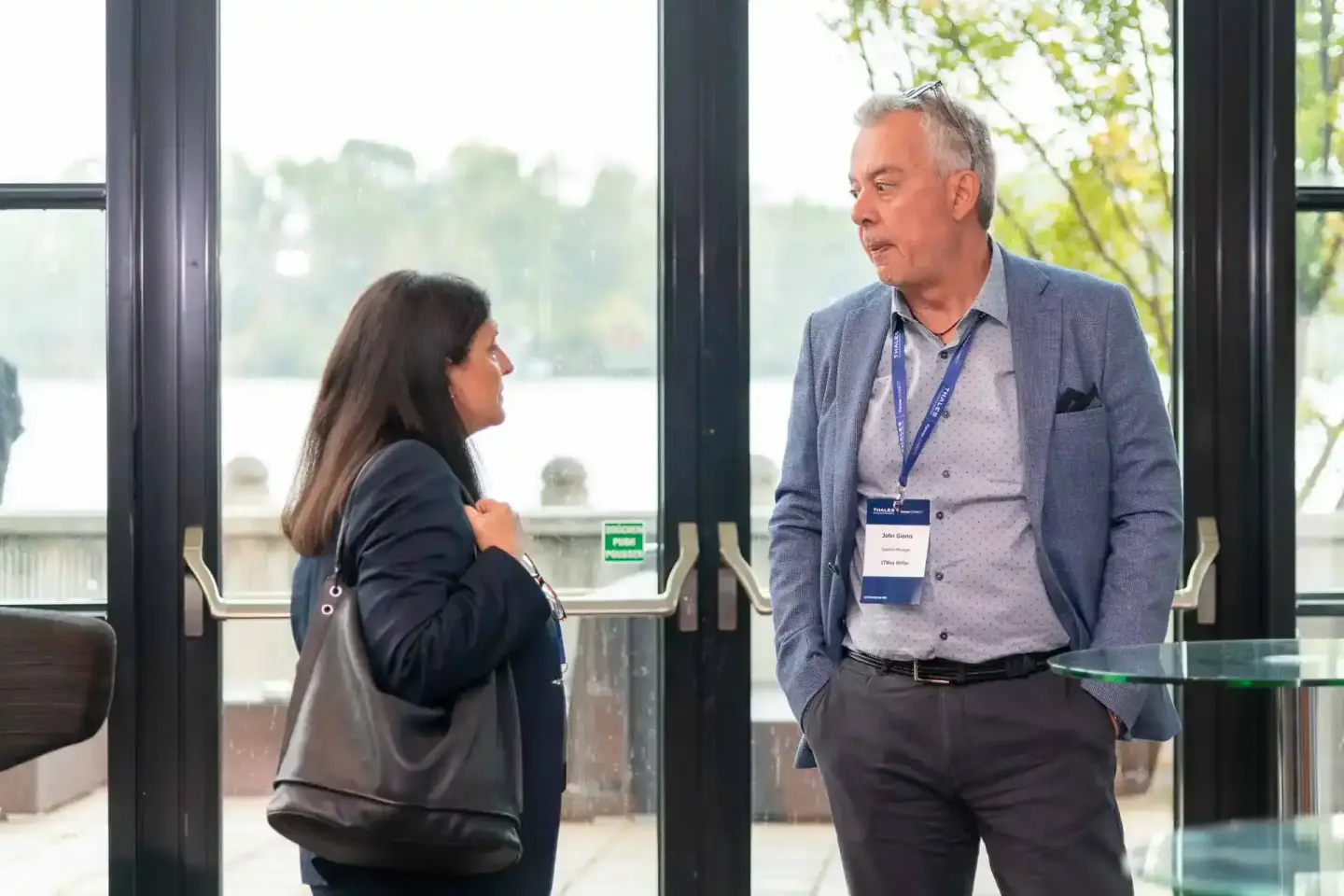Zwei Personen mit Konferenz-Badges im Gespräch vor großen Fenstern mit Blick auf einen See - Eventfotografie Wien