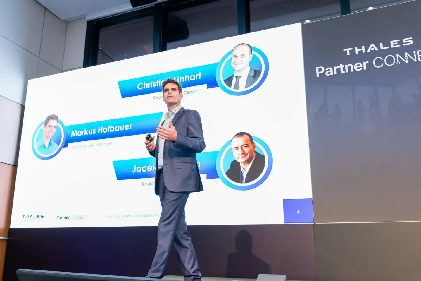 Geschäftsmann präsentiert auf Bühne vor Projektionsscreen mit Mitarbeiterfoto und Thales Partner Connect Logo - Eventfotograf