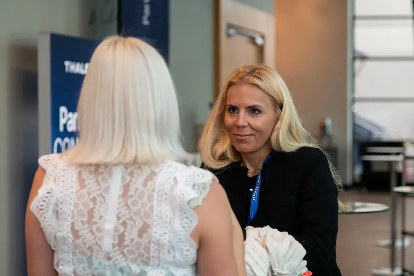 Zwei Frauen im Gespräch bei einem Firmenevent Wien, eine trägt weißes Spitzenkleid, die andere schwarzen Blazer mit Namenssch