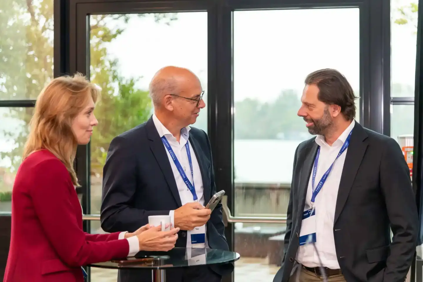 Drei Geschäftsleute in Business-Kleidung mit Namensschildern diskutieren bei einem Firmenevent Wien vor großen Fenstern mit S