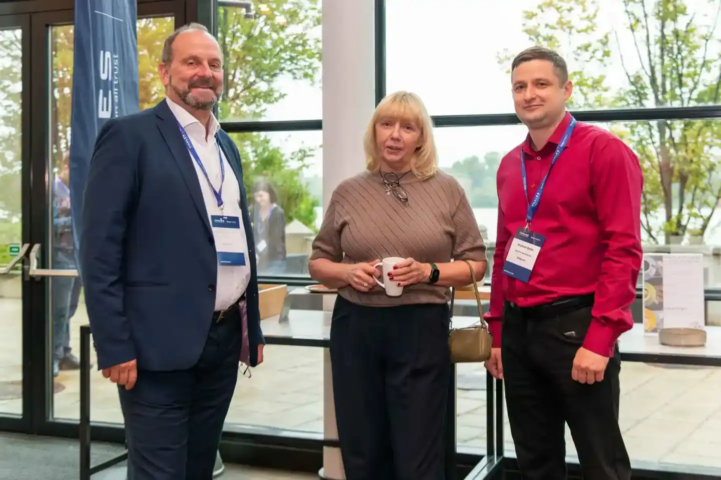 Drei Personen bei einem Firmenevent Wien: Mann im dunklen Anzug, Frau in braunem Pullover mit Kaffeetasse und Mann im roten H