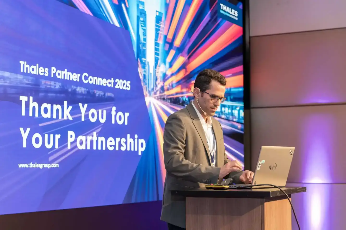 Geschäftsmann präsentiert bei Thales Partner Connect 2025 Event mit Laptop am Rednerpult vor großer Bühne