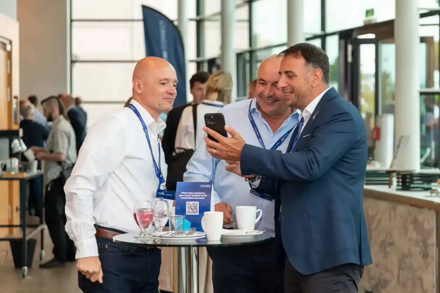 Drei Geschäftsmänner mit Namensschildern bei einem Firmenevent Wien, einer zeigt sein Smartphone, moderner Veranstaltungsort