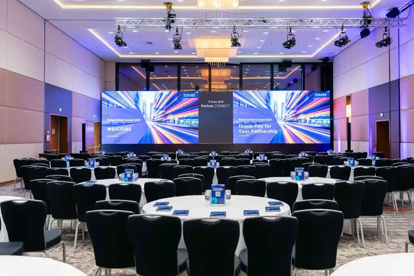 Moderner Konferenzraum mit schwarzen Stühlen an runden Tischen, großer LED-Bildschirm zeigt Thales Partner Connect 2025 Welco