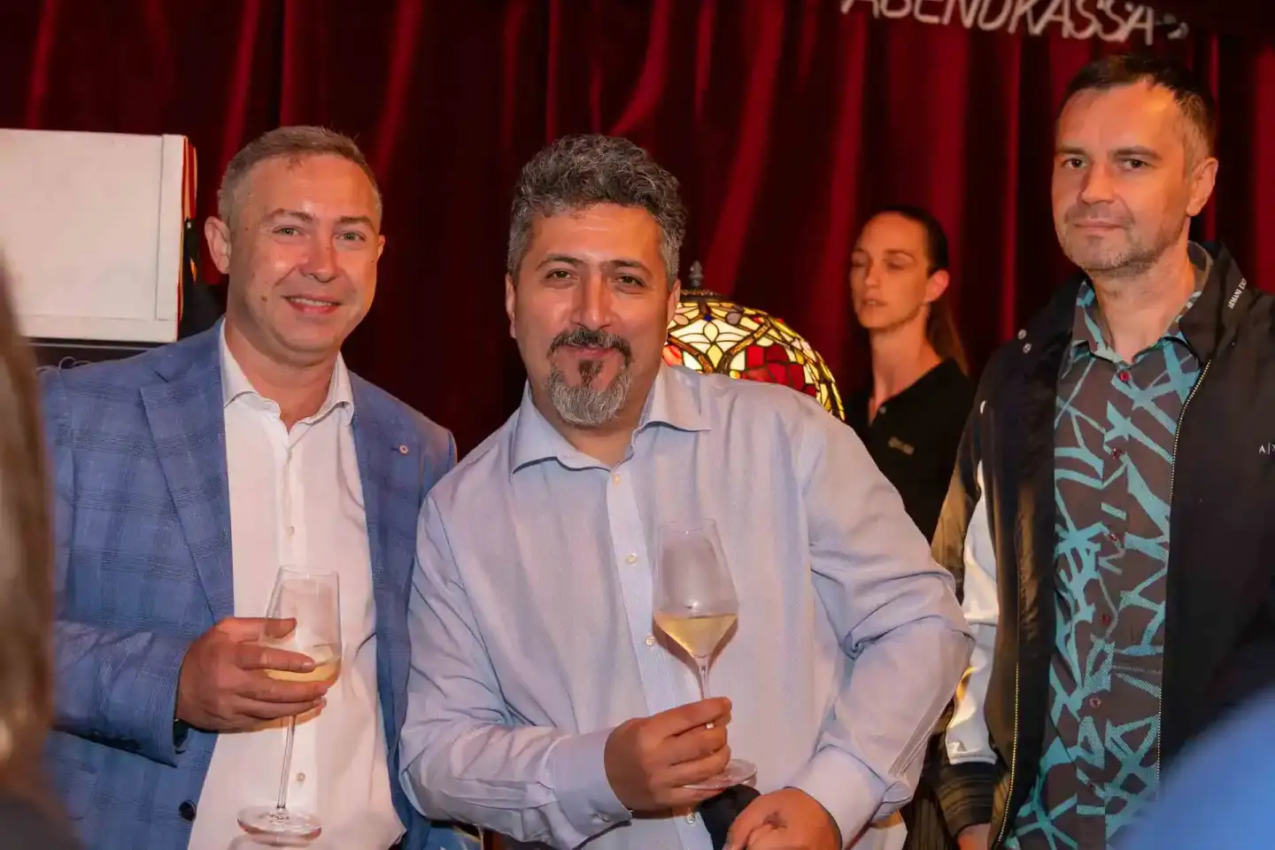Drei Männer in eleganter Kleidung beim Thales Firmenevent Wien, einer hält ein Weinglas, roter Vorhang im Hintergrund