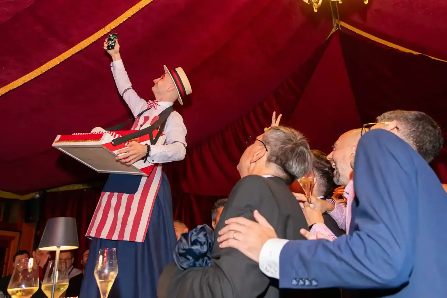 Zirkusartist in rot-weiß gestreiftem Kostüm präsentiert Zaubertrick vor Publikum unter rotem Zelt bei Firmenevent Wien