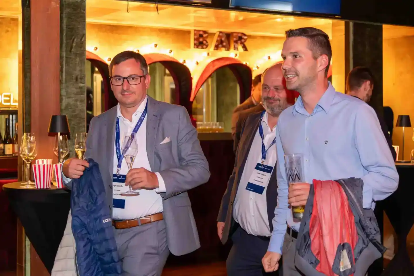Drei Geschäftsmänner mit Namensschildern beim Thales Firmenevent in einer eleganten Bar-Lounge mit goldenem Licht