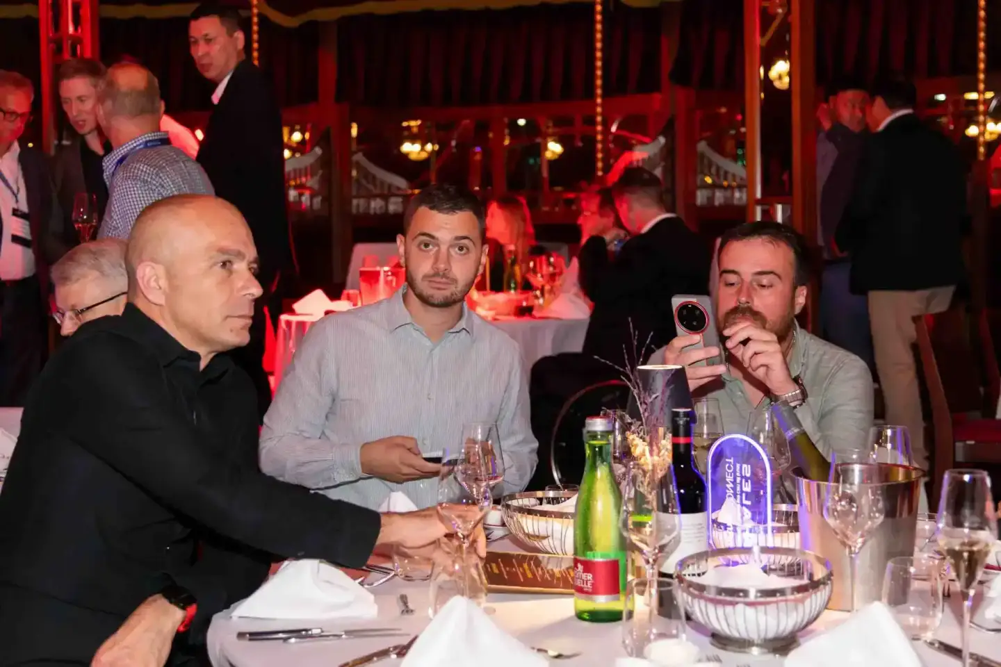 Drei Männer sitzen an einem festlich gedeckten Tisch bei einem Firmenevent Wien, im Hintergrund rote Beleuchtung und weitere 