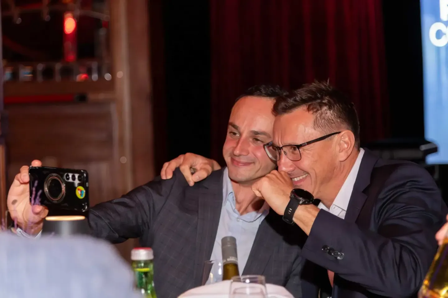 Zwei Geschäftsmänner machen ein Selfie bei einem Firmenevent Wien, einer hält eine Kamera, beide lachen