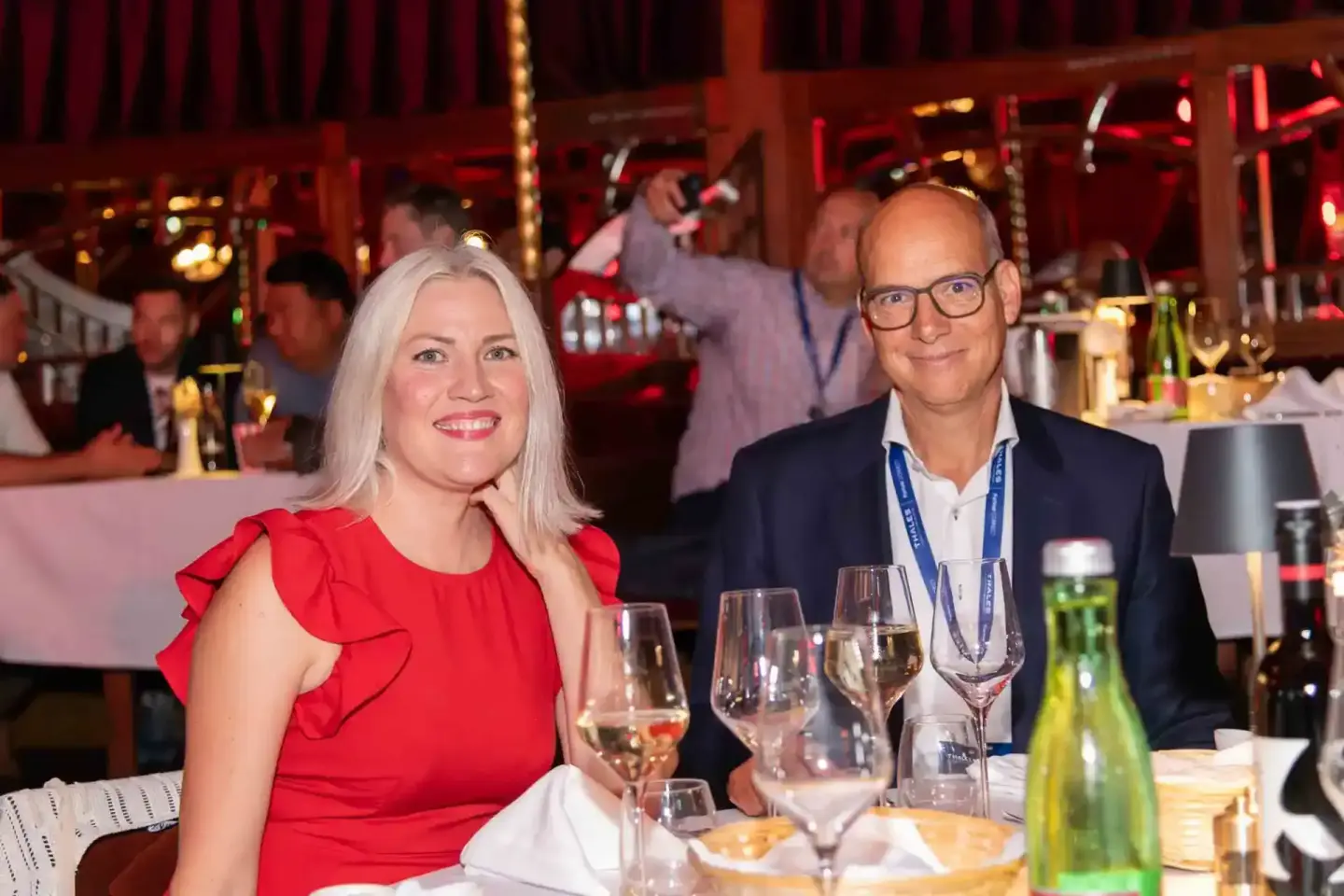 Zwei elegante Gäste beim Thales Firmenevent in Wien sitzen am Tisch mit Weingläsern und lächeln in die Kamera