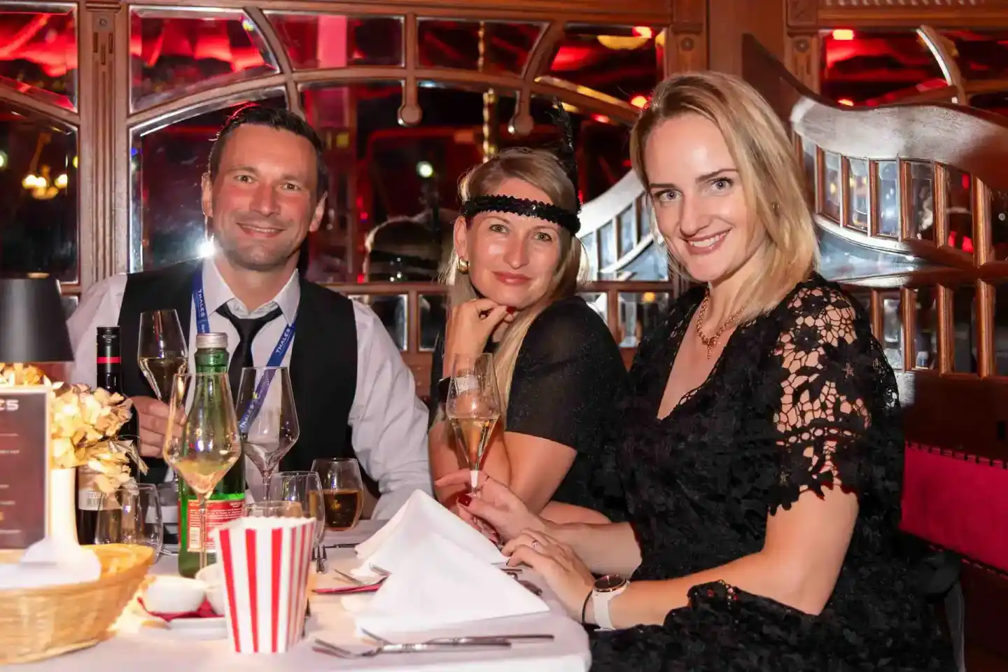 Drei elegant gekleidete Personen bei einem Firmenevent Wien an einem Tisch mit Champagner und Popcorn, vor roten Fenstern ein