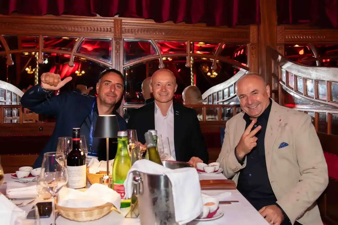 Drei Geschäftsmänner beim Firmenevent Wien an einem eleganten Restauranttisch mit Wein und Gedeck, im Hintergrund rotes Theat