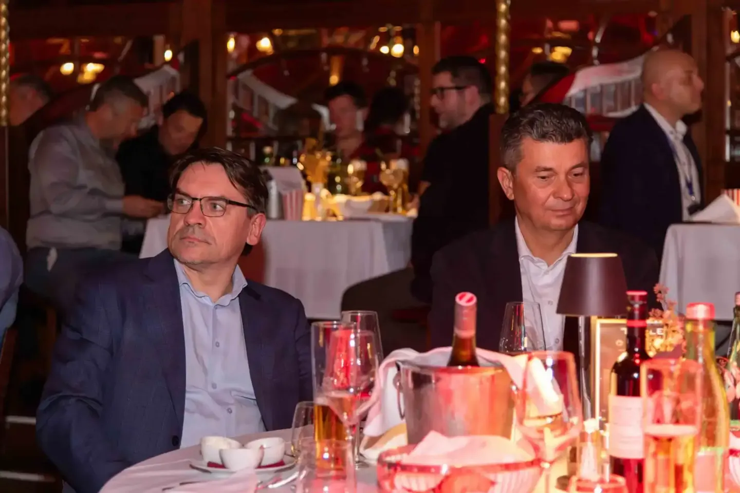 Zwei Geschäftsmänner sitzen beim Firmenevent Wien an einem gedeckten Tisch mit Getränken und beobachten eine Veranstaltung.