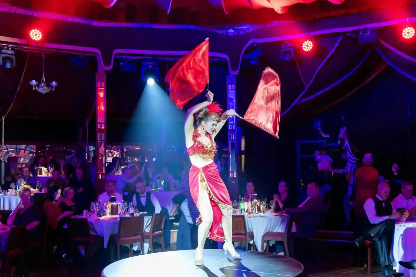 Performerin in rotem Kostüm mit Federn tanzt auf Bühne bei Firmenevent Wien, Publikum sitzt an Tischen mit Getränken
