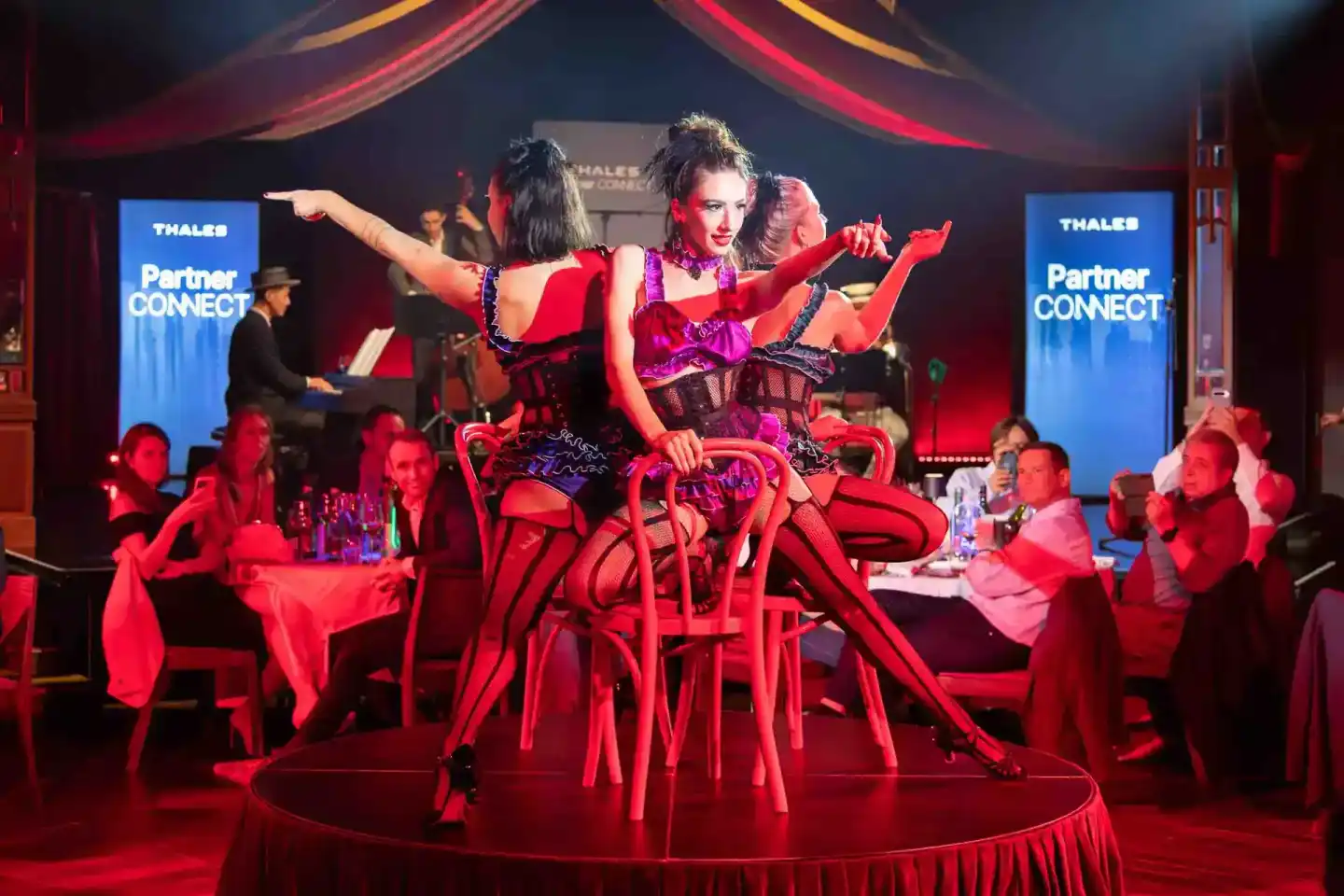 Performerin in rotem Kostüm tanzt auf Bühne beim Thales Partner Connect Event, umgeben von Gästen an roten Tischen - Eventfot