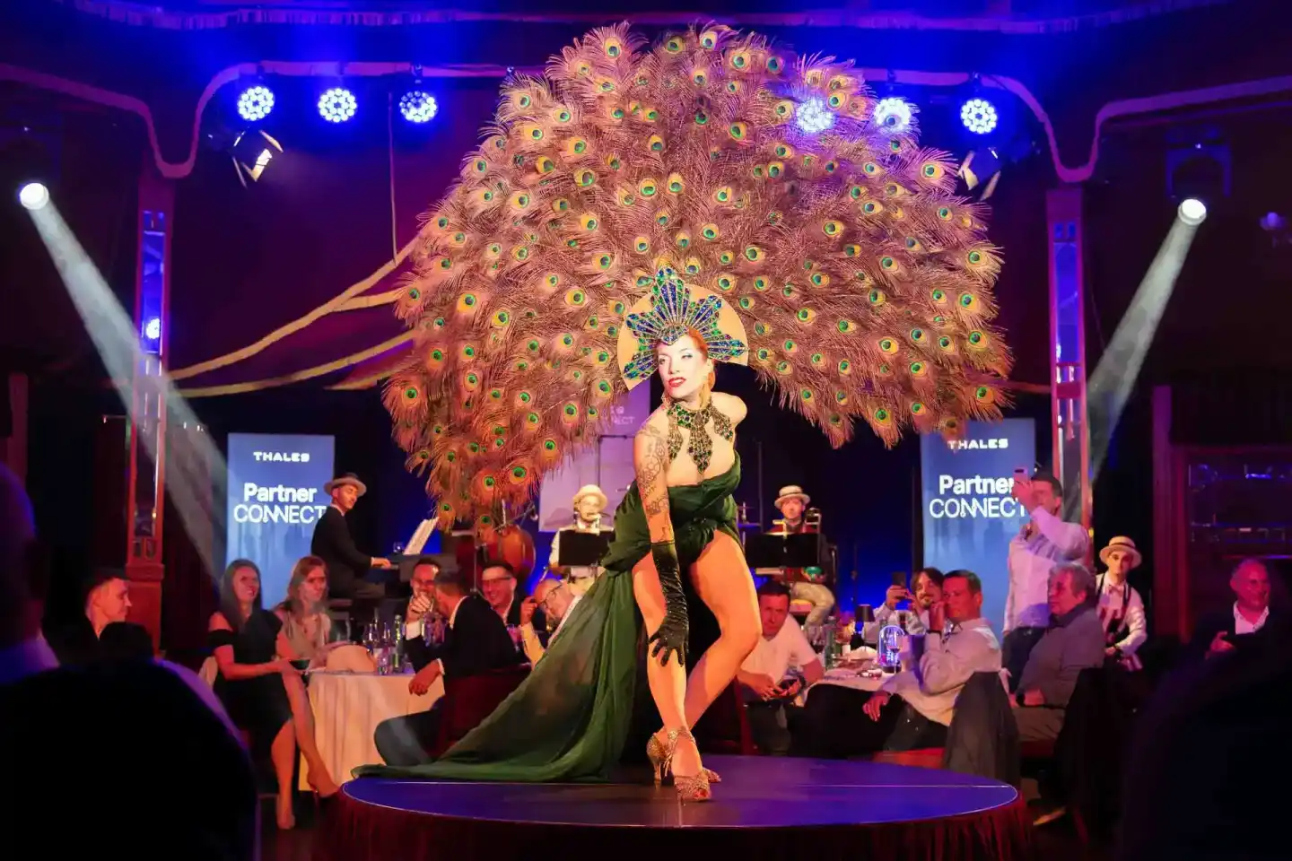 Performerin in grünem Kostüm mit riesigem orangefarbenen Pfauenfeder-Kostüm auf der Bühne bei Thales Partner Connect Event