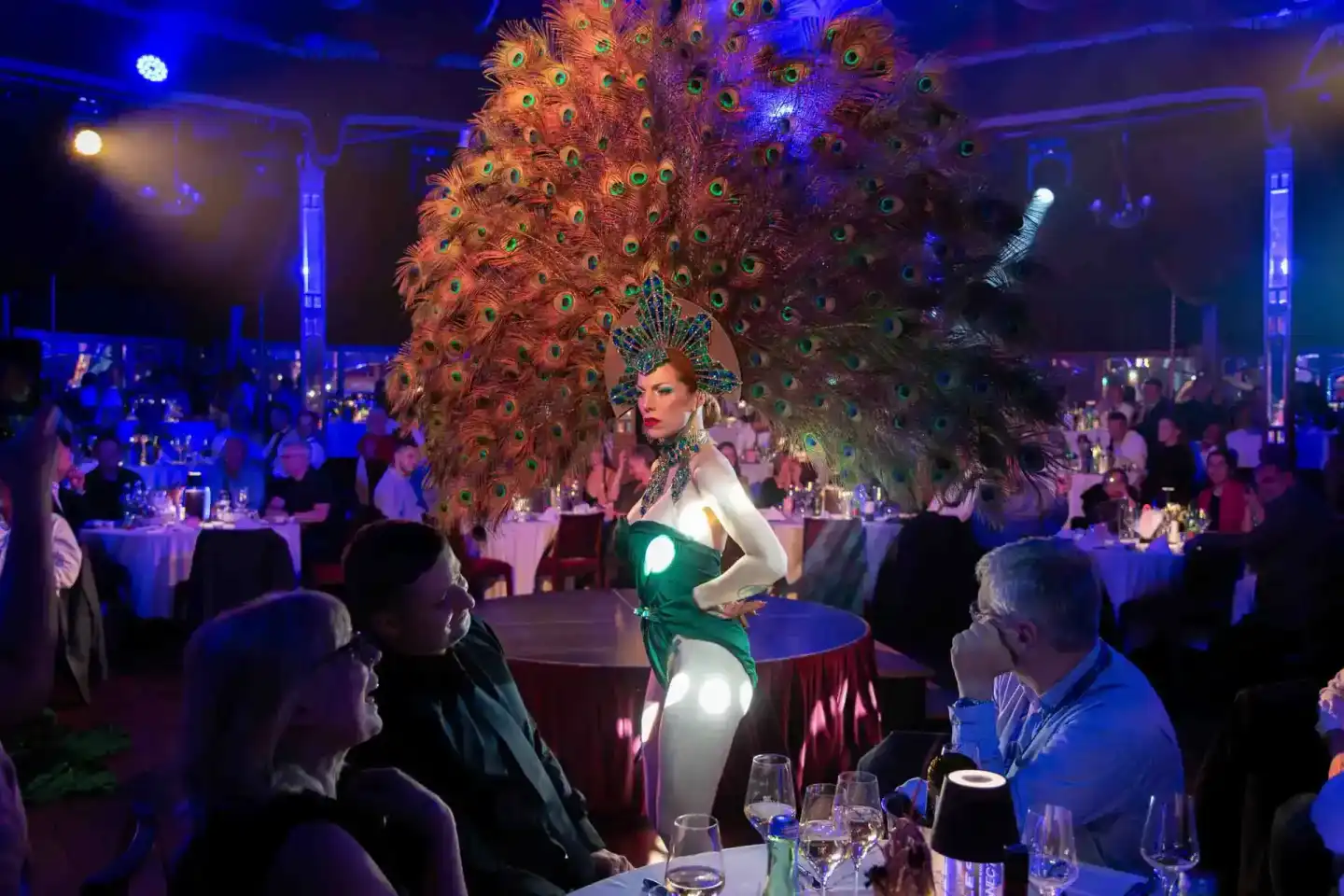 Elegante Performerin im grünen Kleid vor riesigem Pfauenfeder-Kostüm bei Gala-Event, Eventfotograf Wien dokumentiert Bühnenau