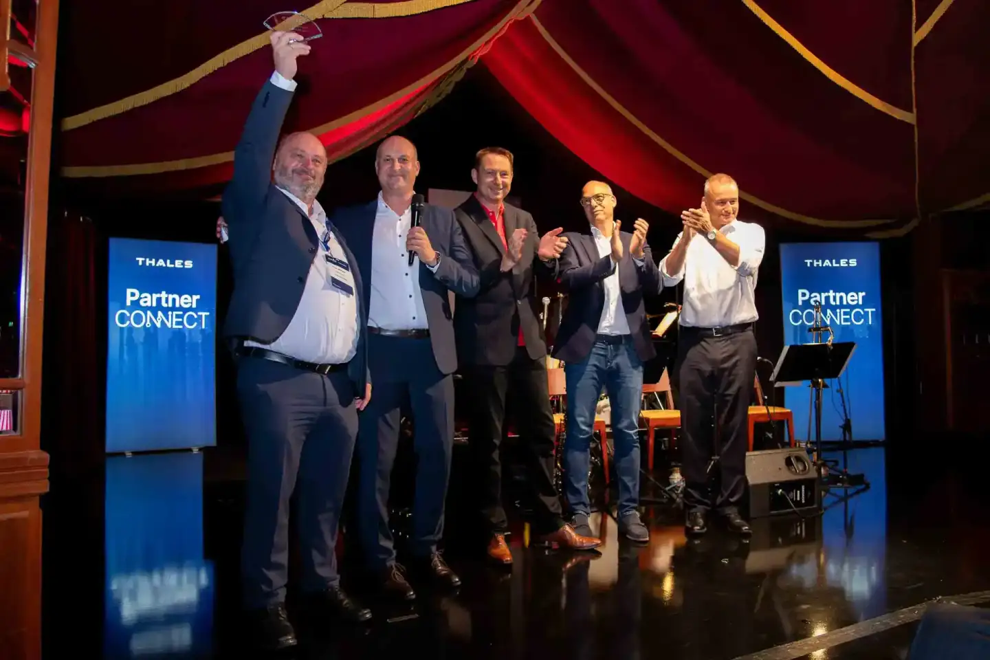 Fünf Geschäftsmänner auf der Bühne der Thales Partner Connect Veranstaltung, einer hebt triumphierend die Faust. Professionel