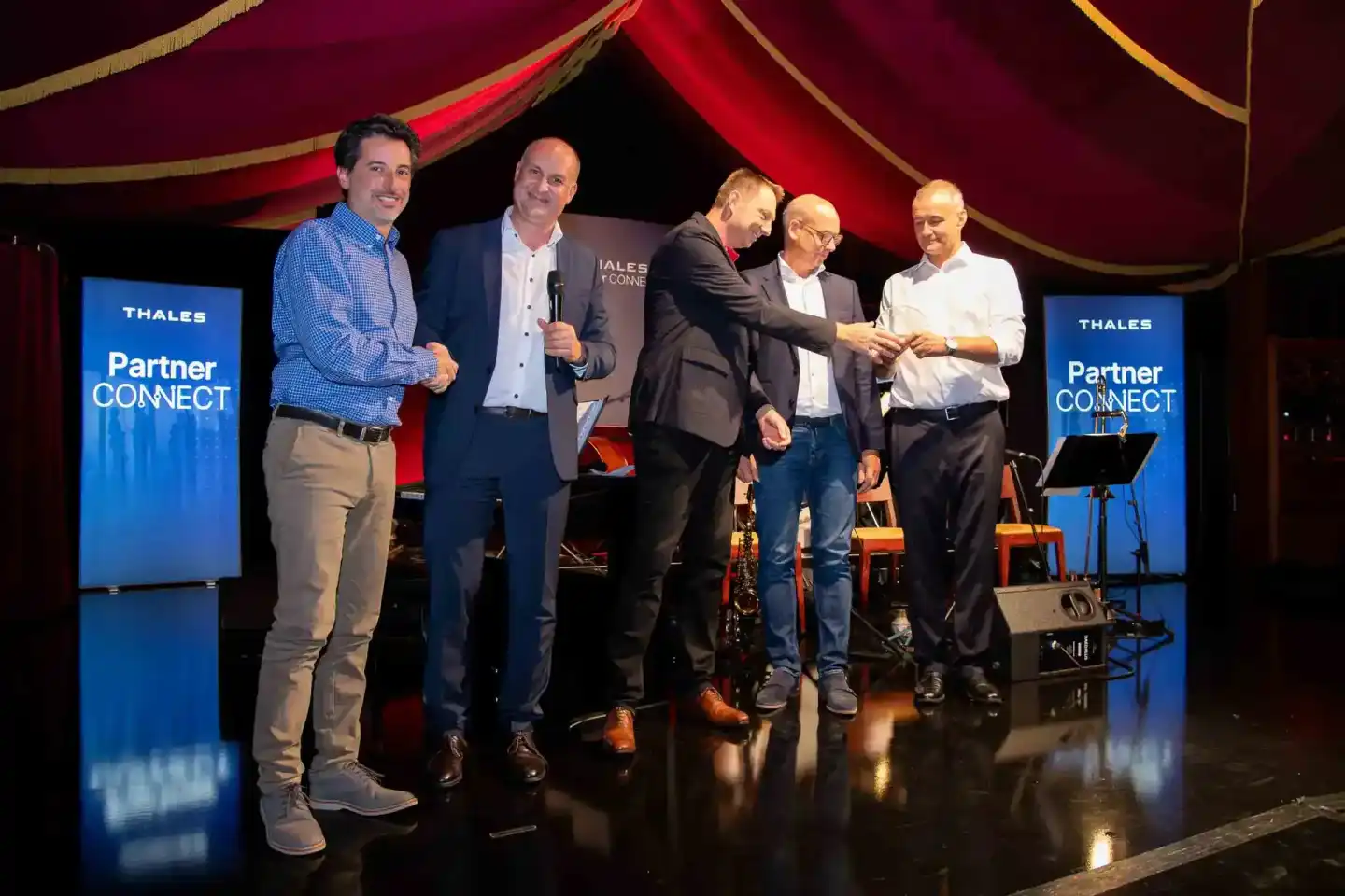 Fünf Geschäftsmänner auf der Bühne des Thales Partner Connect Events in Wien, einer moderiert mit Mikrofon