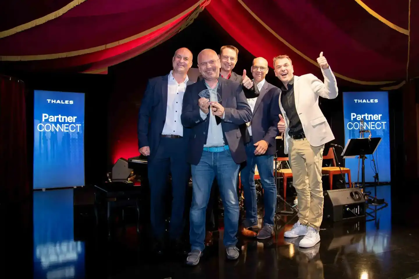 Fünf Geschäftsmänner auf der Bühne des Thales Partner Connect Events, lächelnd mit Daumen nach oben bei einer Veranstaltungsf