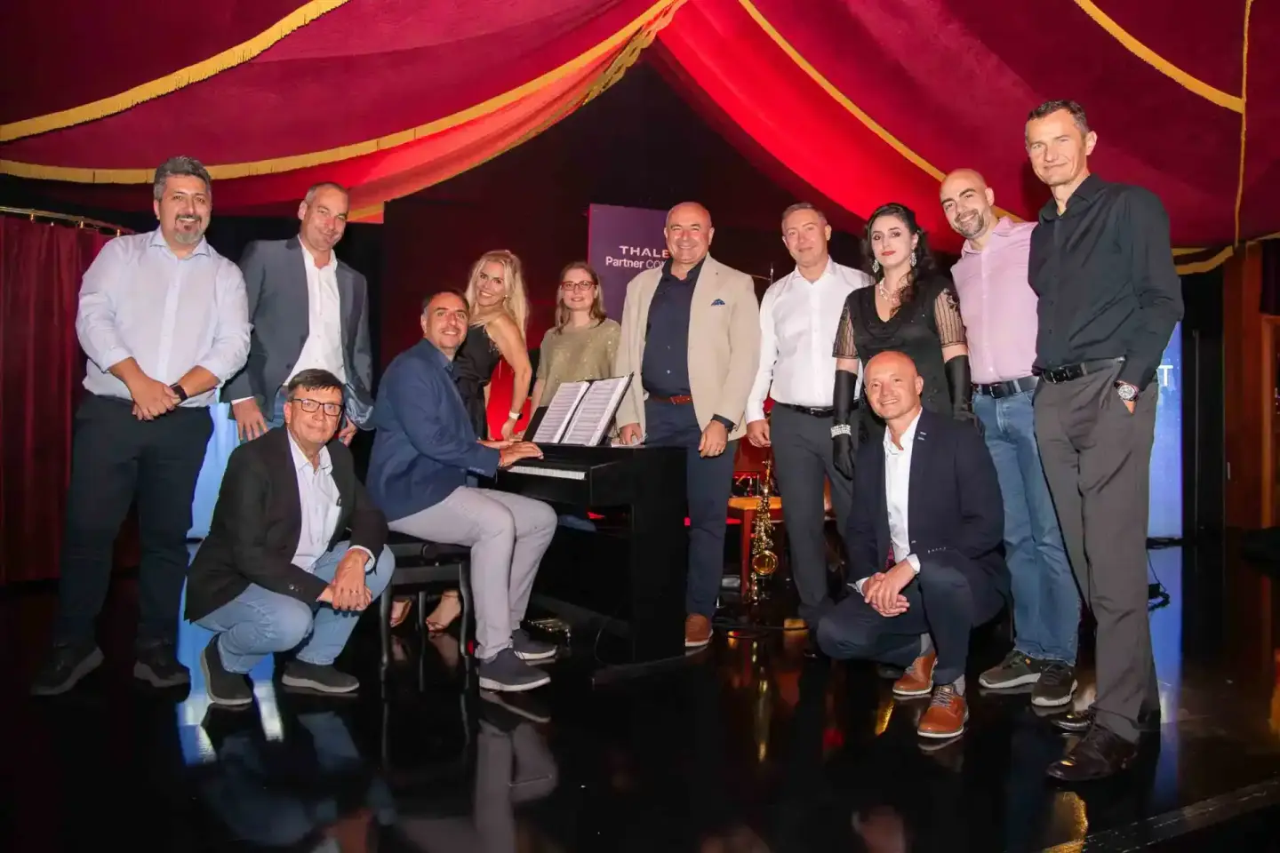 Gruppenfoto von etwa 12 Personen bei einem Thales Firmenevent in Wien, stehend und kniend um ein Klavier, elegant gekleidet v