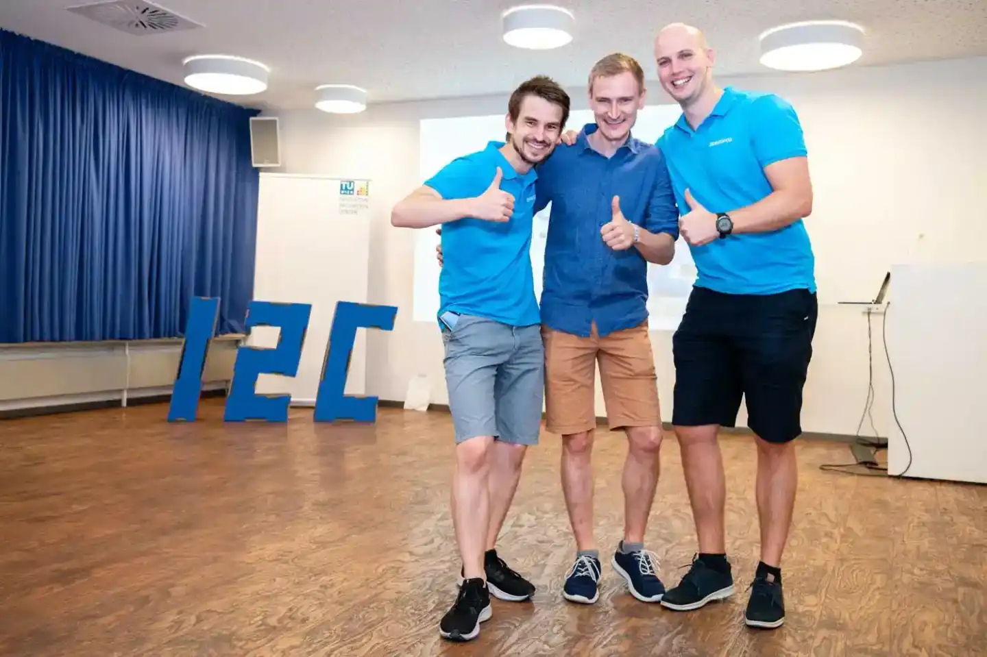 Drei lachende Männer in blauen Polo-Shirts geben Daumen nach oben vor großen IFT-Buchstaben bei einem Firmenevent Wien
