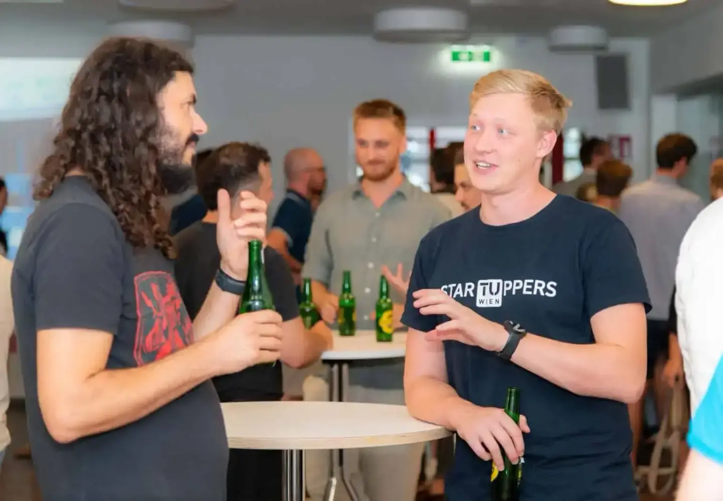 Zwei Männer unterhalten sich auf einem Firmenevent Wien mit Bierflaschen in der Hand, weitere Gäste im Hintergrund