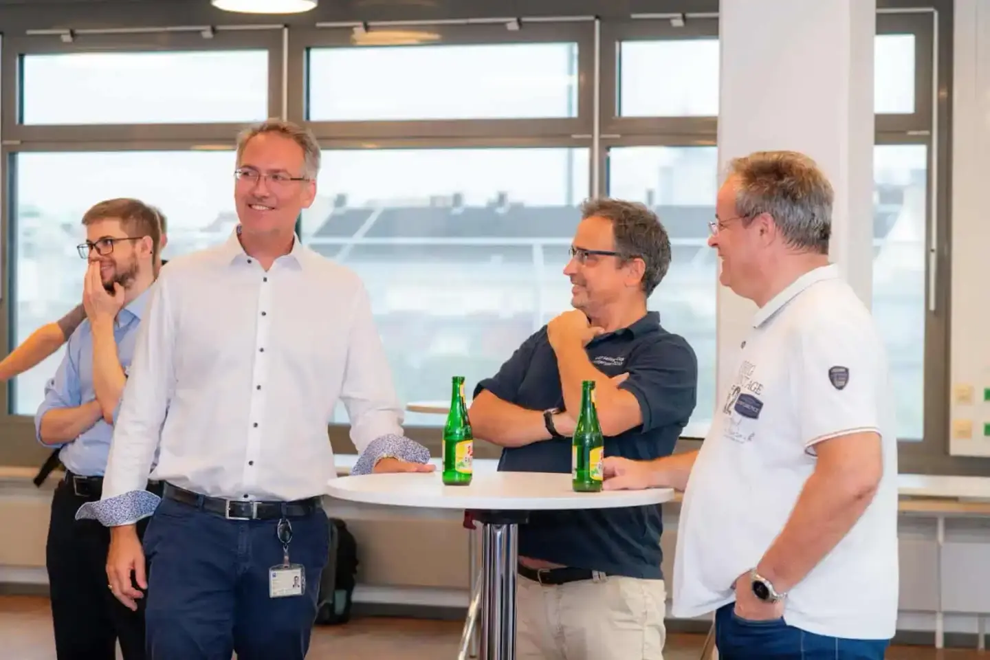 Vier Männer unterhalten sich bei einem Firmenevent Wien an einem Stehtisch mit Getränken, moderne Bürokulisse mit großen Fens