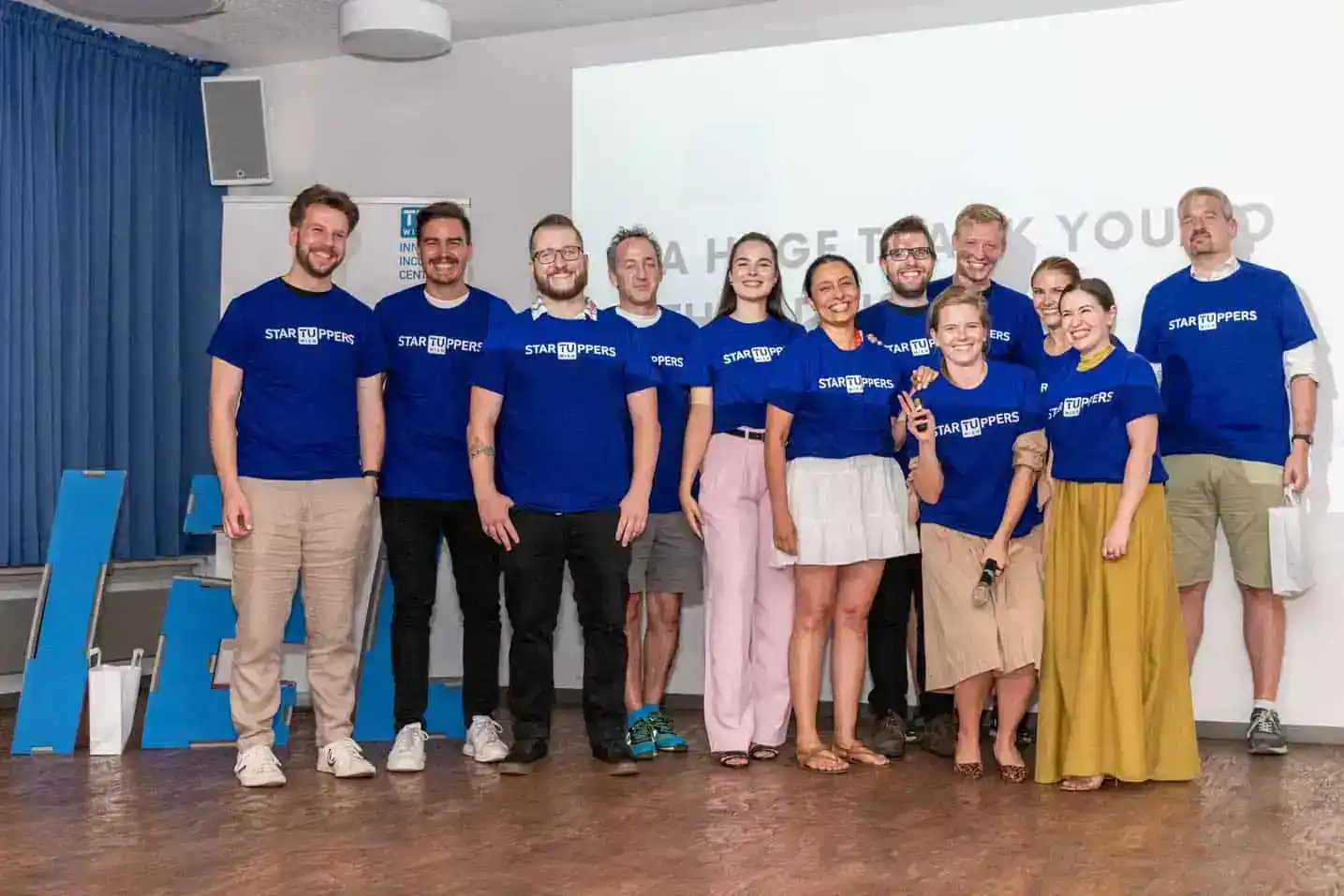 Team der TU Wien in blauen StarUppers-Shirts bei einem Firmenevent, lachend und zusammenstehend vor Präsentationswand
