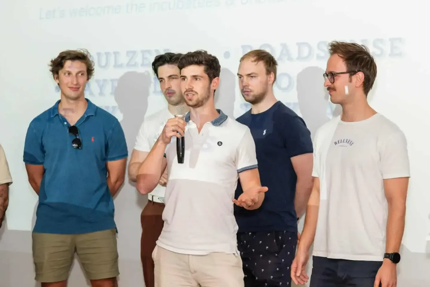 Fünf junge Männer präsentieren sich auf einer Bühne während eines Firmenevents. Ein Sprecher hält ein Mikrofon, die anderen s