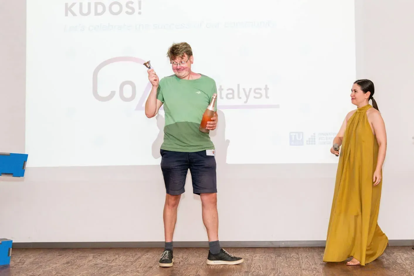 Mann in grünem Shirt mit Sektflasche präsentiert Preis auf TU Wien Veranstaltung, Eventfotograf dokumentiert Moment